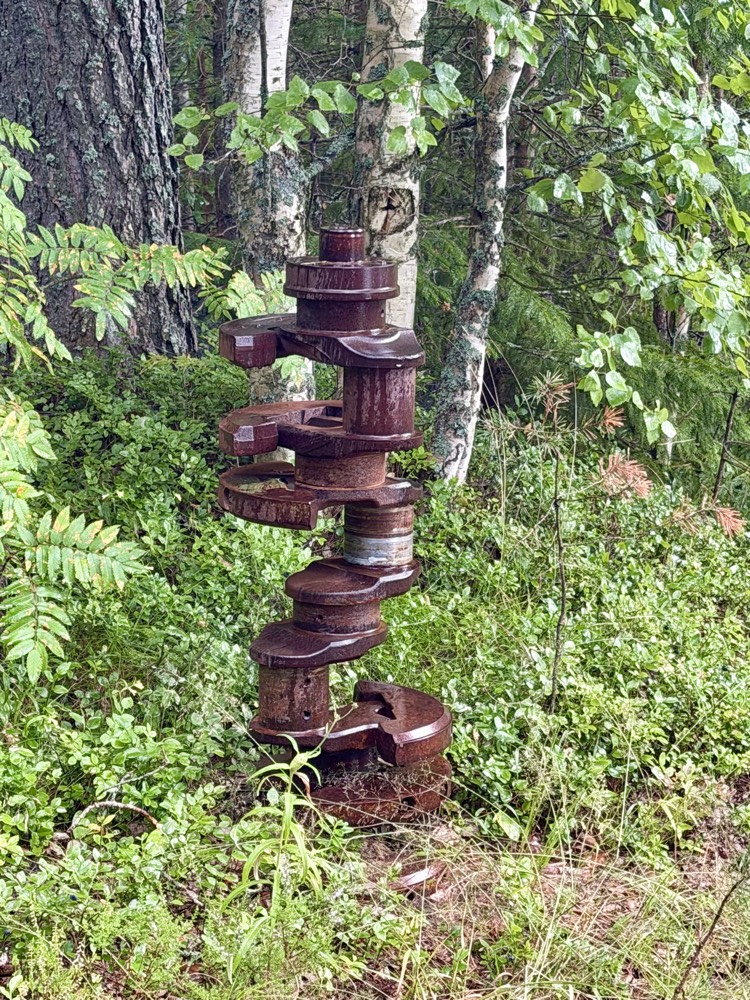 Skulptur aus gestapelten verrosteten Maschinenteilen steht zwischen Birken im Unterholz