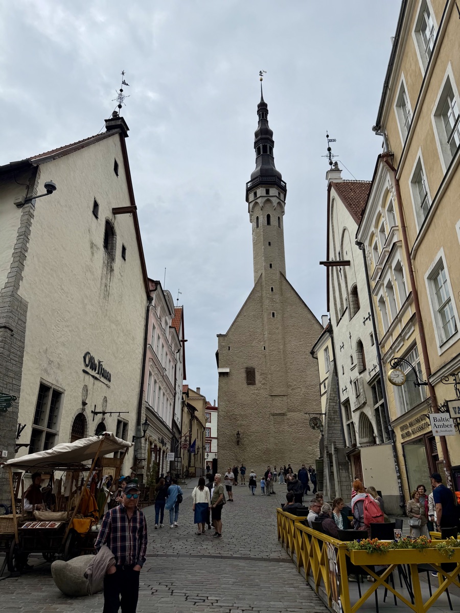Schlanker Kirchturm mit dunkler Spitze am Ende einer Gasse, davor Menschen an einem Café