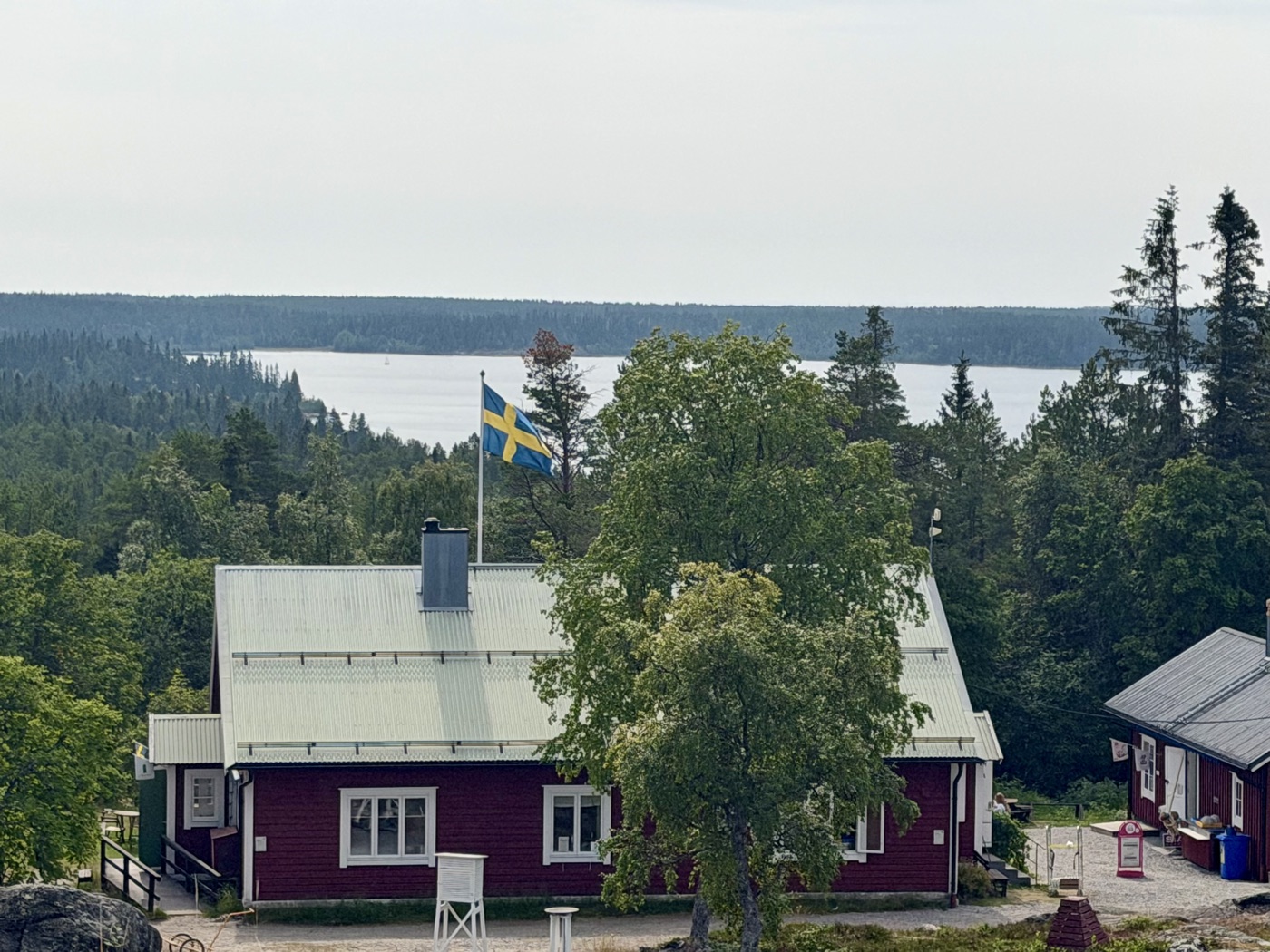 Rotes Schwedenhaus mit schwedischer Flagge, dahinter der Wald und das Meer