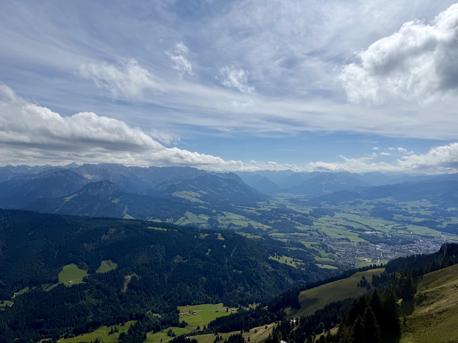 Blick vom Gipfel in ein Tal mit einer Stadt und dahinter Bergketten unter Wolken