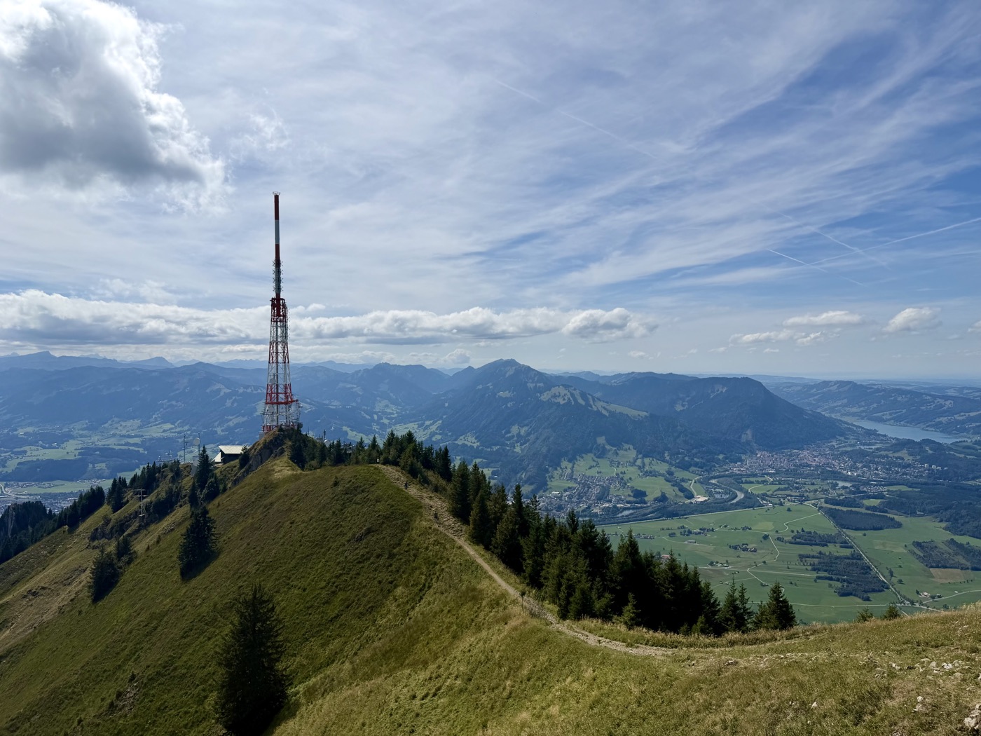 Grasbewachsener Grat mit rot-weißem Sendemast, dahinter Berge und ein See