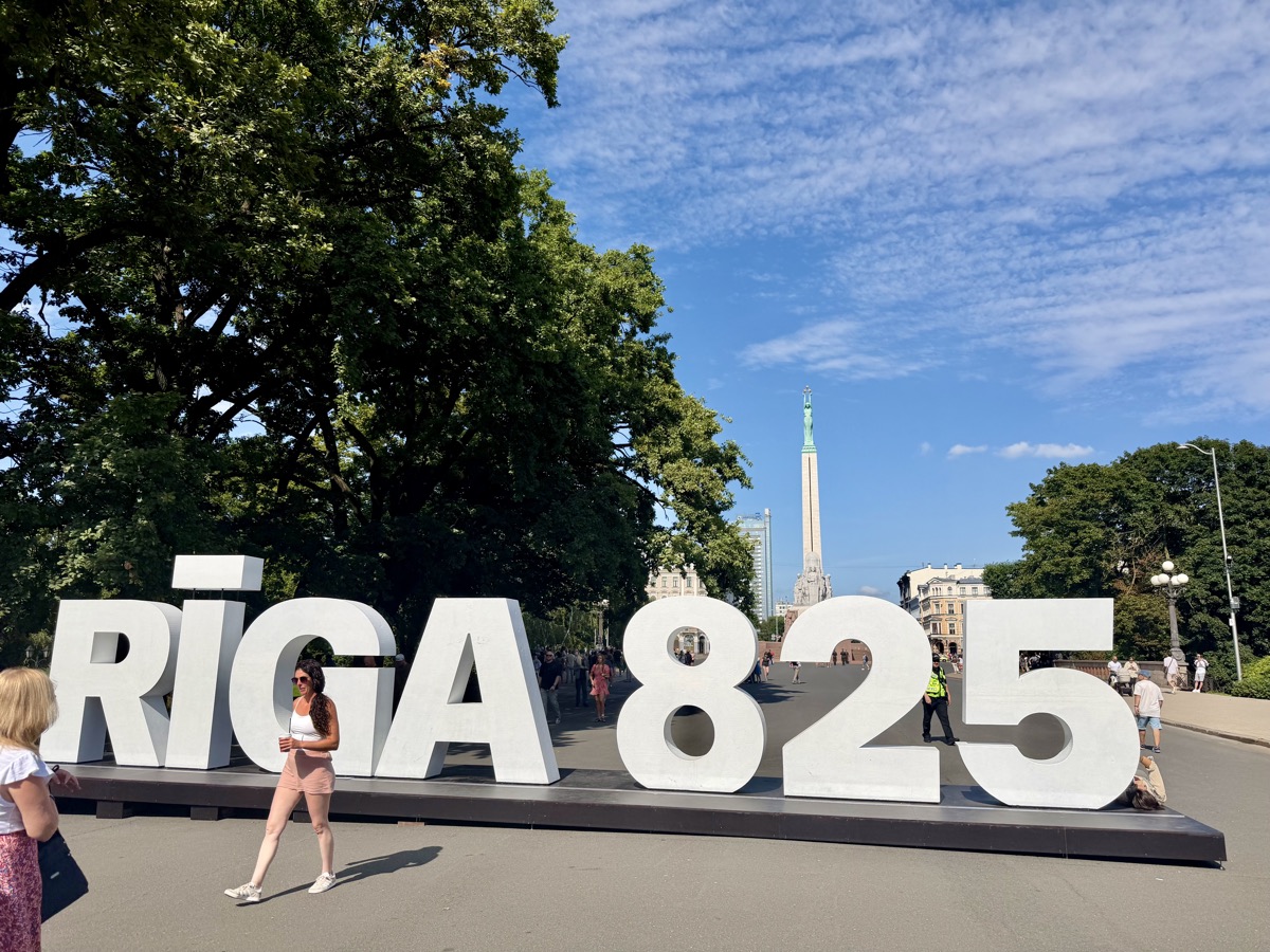 Große weiße Buchstaben Riga 825 auf einer Bühne, dahinter das Freiheitsdenkmal