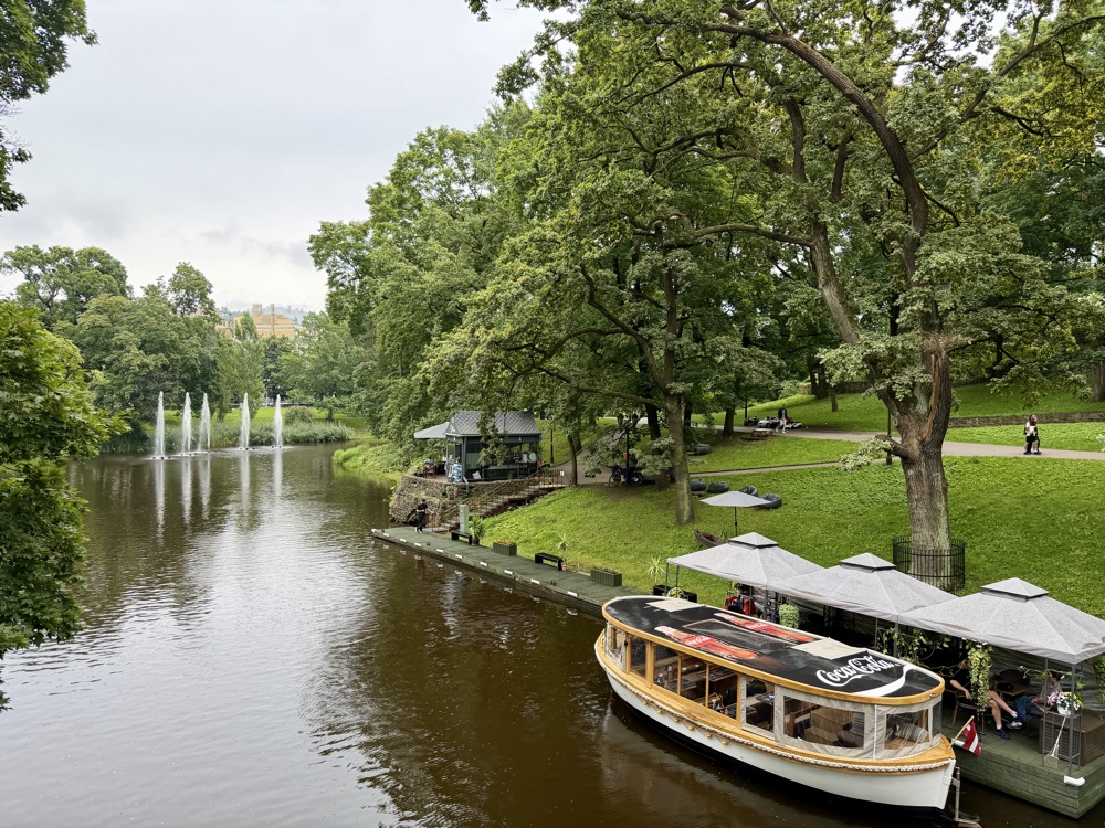 Ruhiger Stadtkanal mit Ausflugsboot zwischen grünen Bäumen, im Hintergrund Fontänen