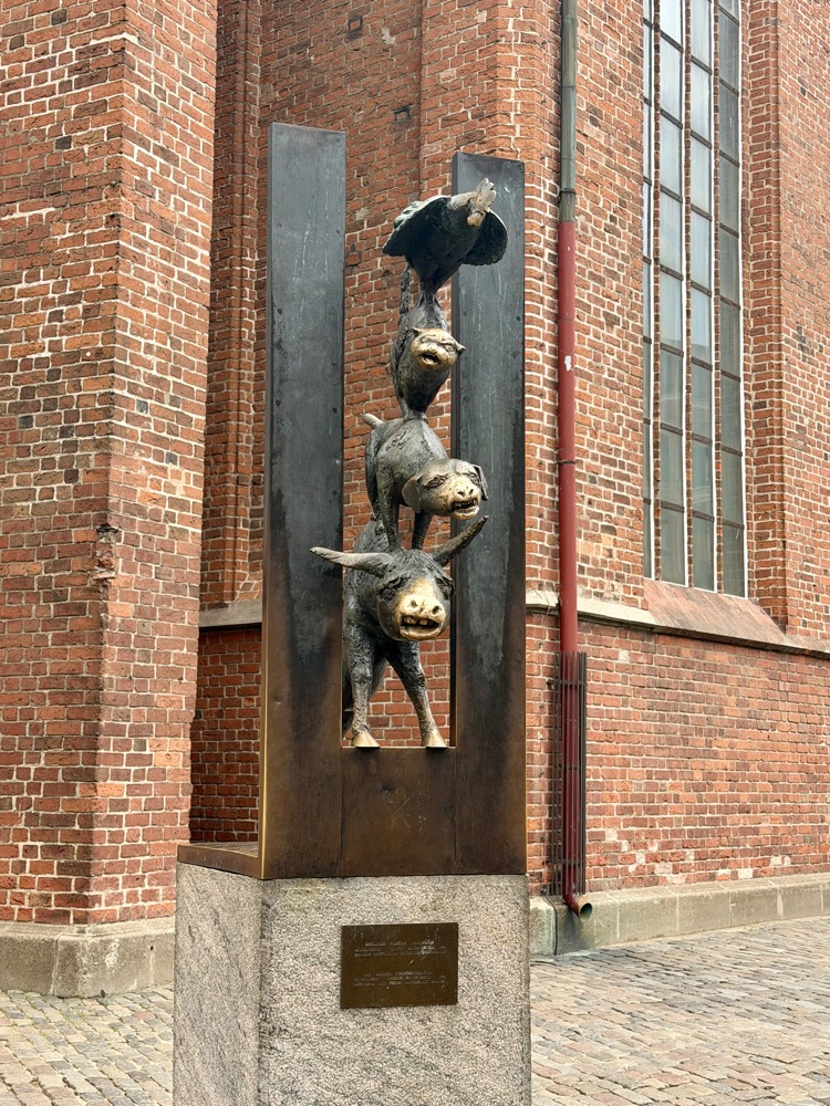 Bronzeskulptur von Esel, Hund, Katze und Hahn übereinander vor einer Backsteinwand