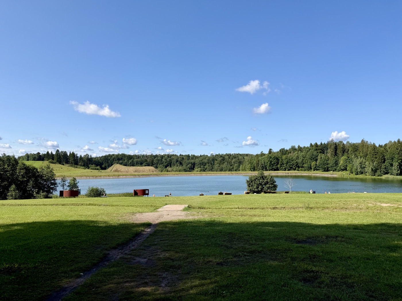 Blick über eine Wiese auf einen kleinen See, an dessen Ufer zwei rote Saunahütten stehen
