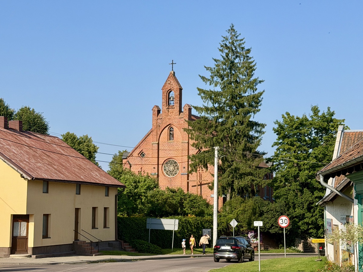Rote Backsteinkirche mit spitzem Turm hinter Bäumen an einer Dorfstraße
