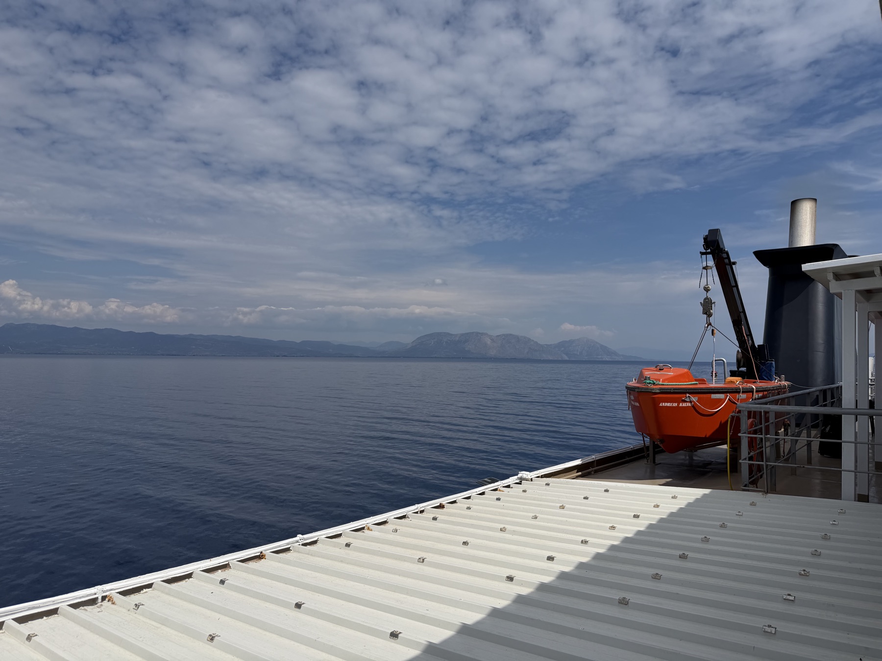 Blick vom Fährdeck mit orangem Rettungsboot über das ruhige Meer auf eine Bergkette