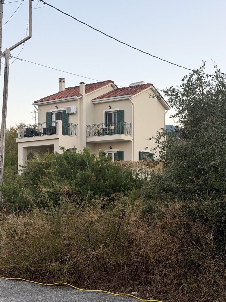 Zweistöckiges helles Haus mit Balkon und roten Dachziegeln hinter Büschen