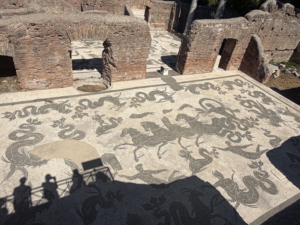 Schwarz-weißes Bodenmosaik mit Meerwesen in einem römischen Badehaus, im Vordergrund die Schatten der Besucher