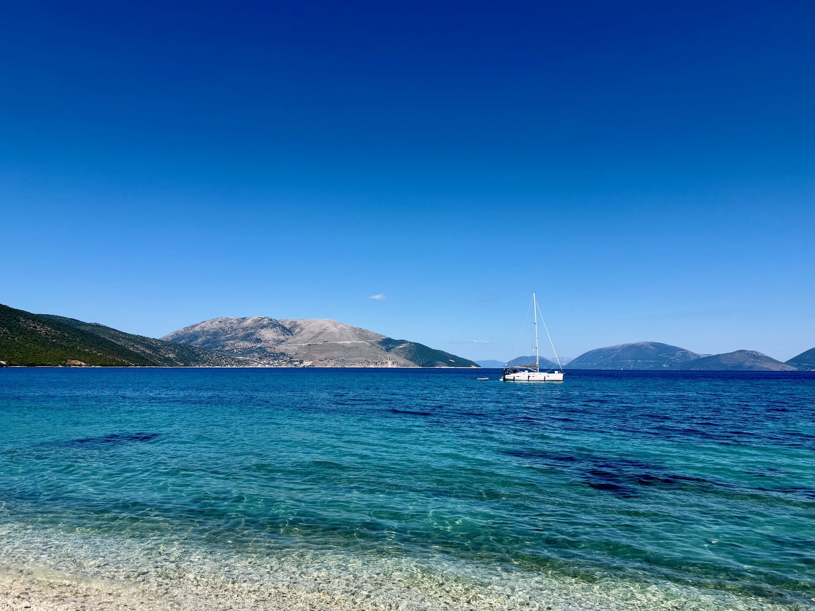 Kieselstrand und tiefblaues Wasser, im Hintergrund ein Segelboot und Berge